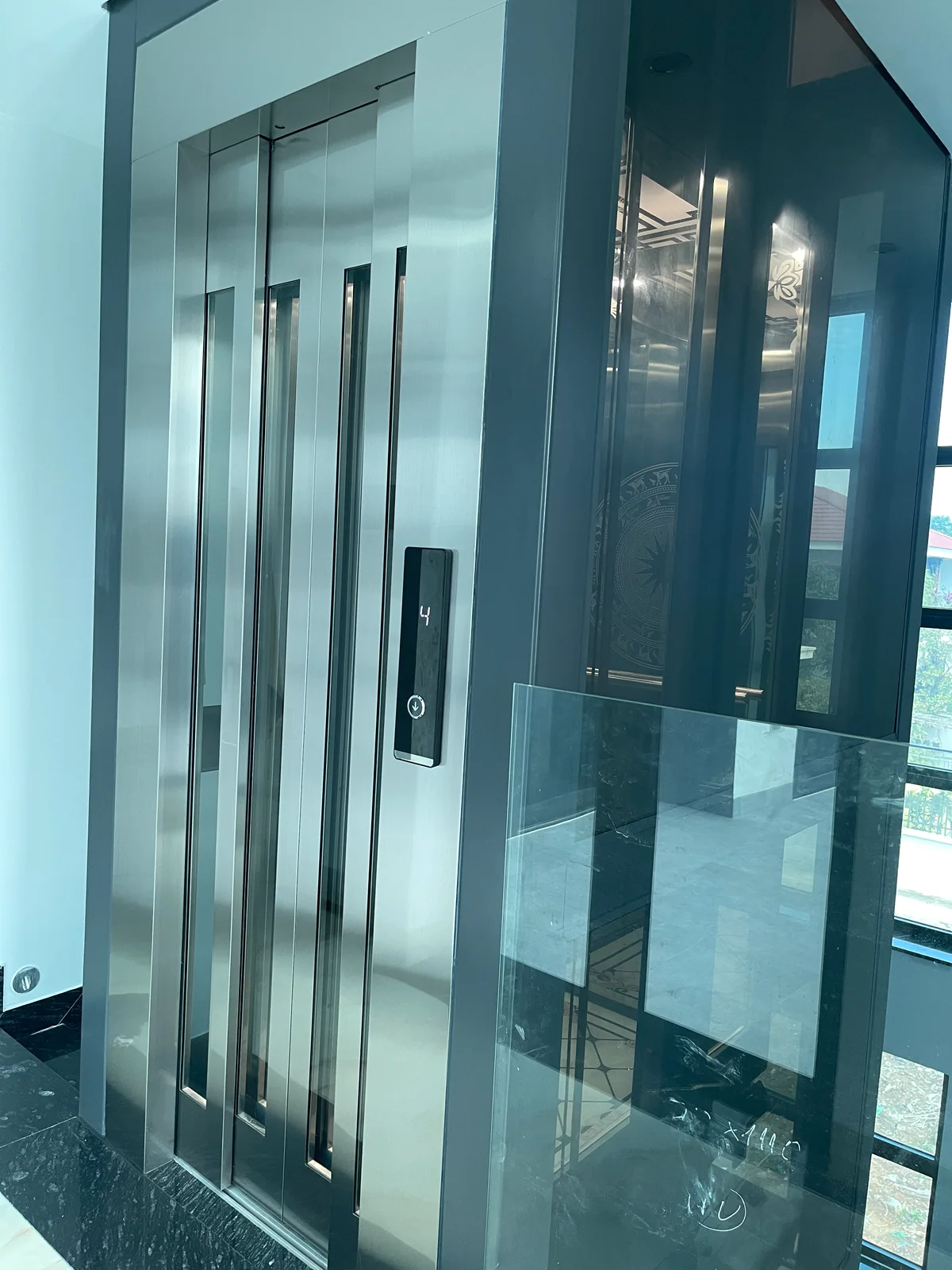 Thang kính khung xám với cửa inox có hai ô kính dọc.