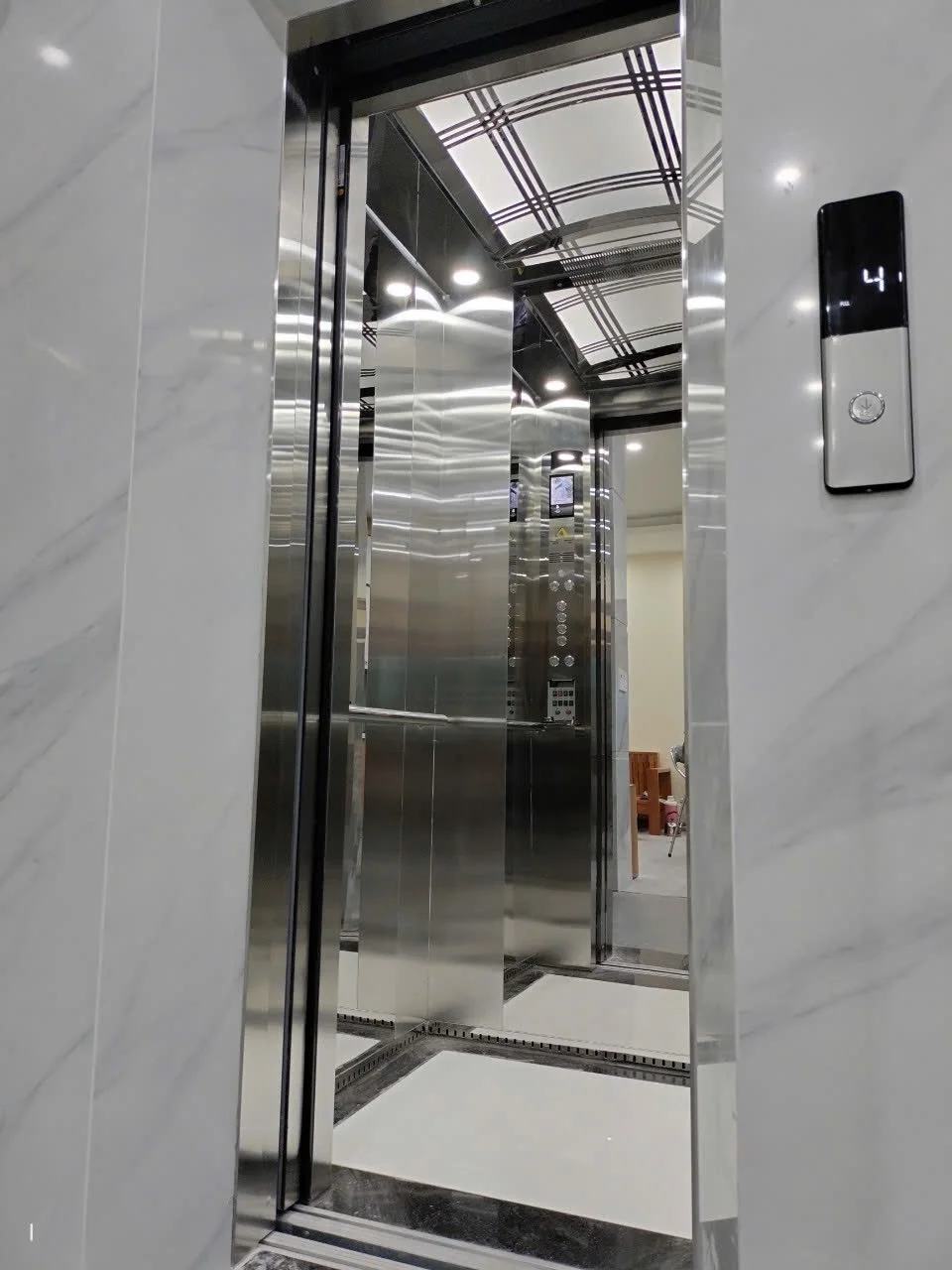 Cửa thang mở trong khung đá trắng, cabin sáng với sàn viền đen.