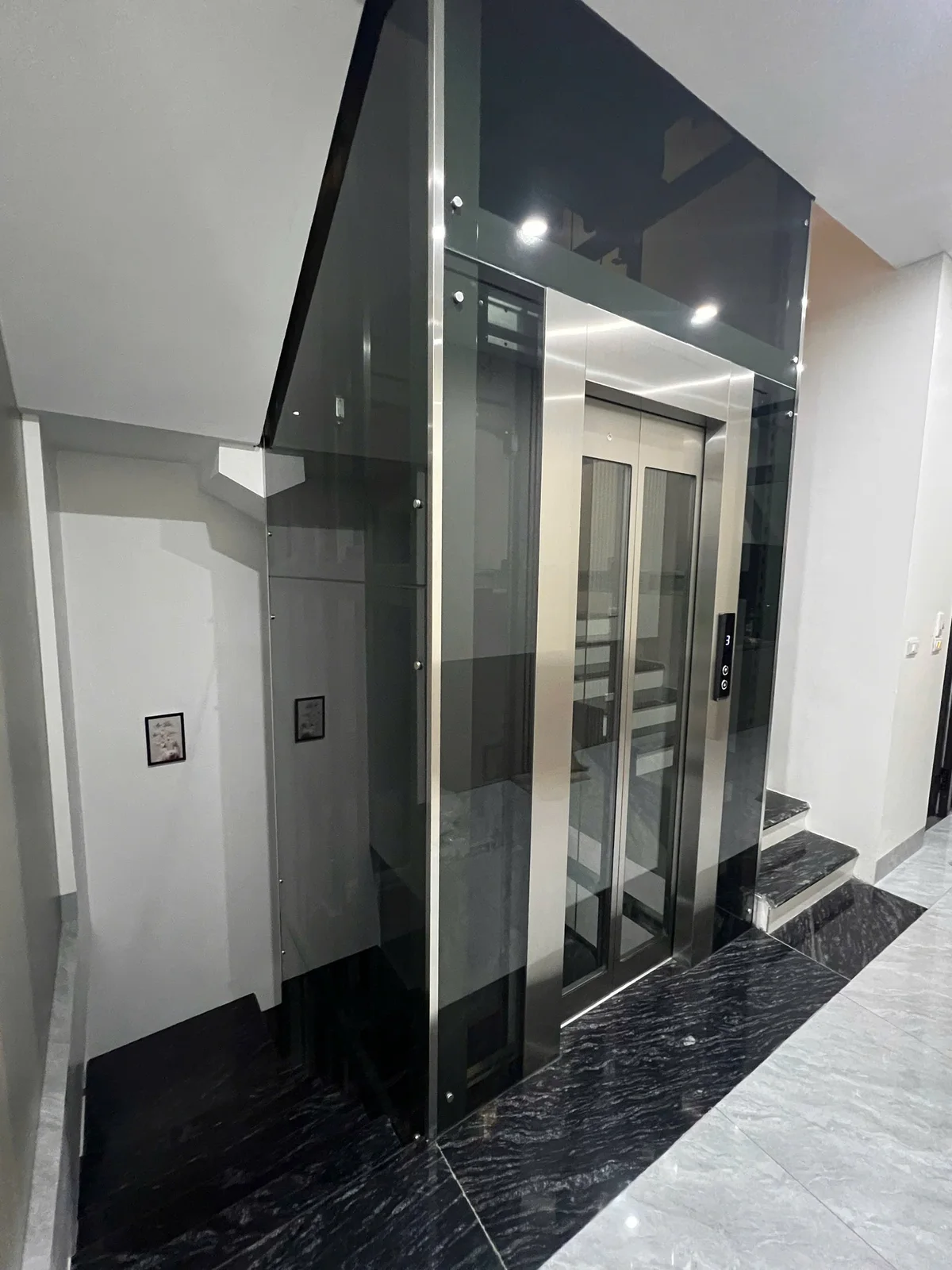 Thang kính có cửa inox và ô kính dọc, nằm cạnh cầu thang bộ.