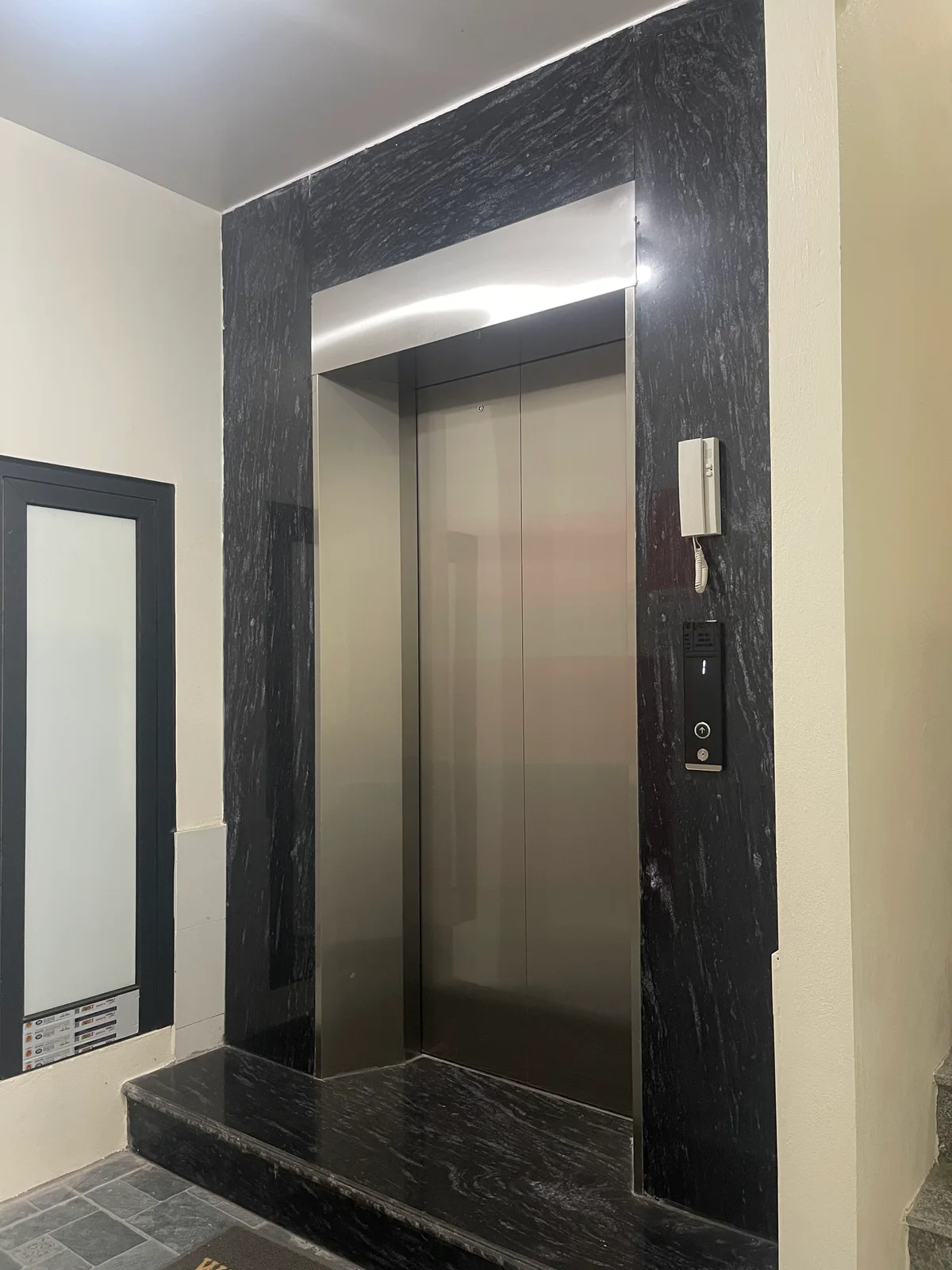 Cửa thang inox đóng, khung đá đen và bậc đá phía trước.