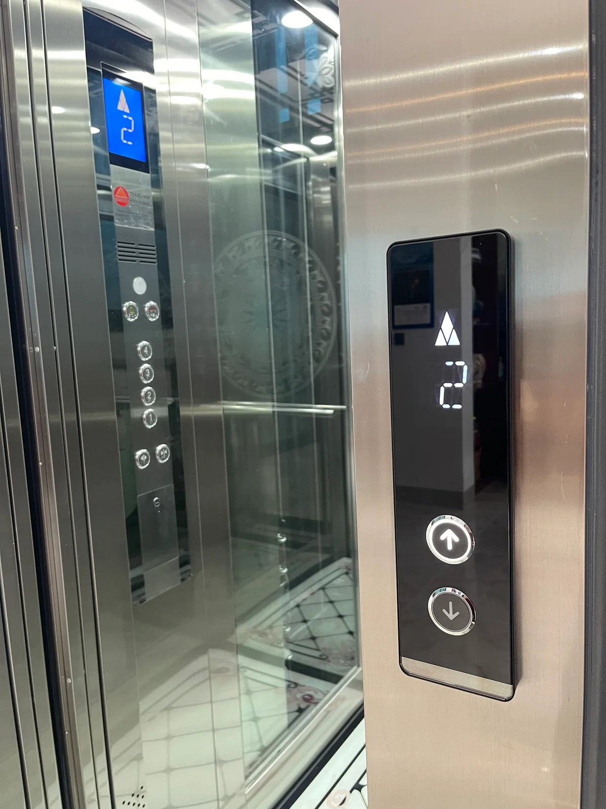 Bảng gọi tầng có màn hình số và nút mũi tên, cạnh cửa cabin mở.
